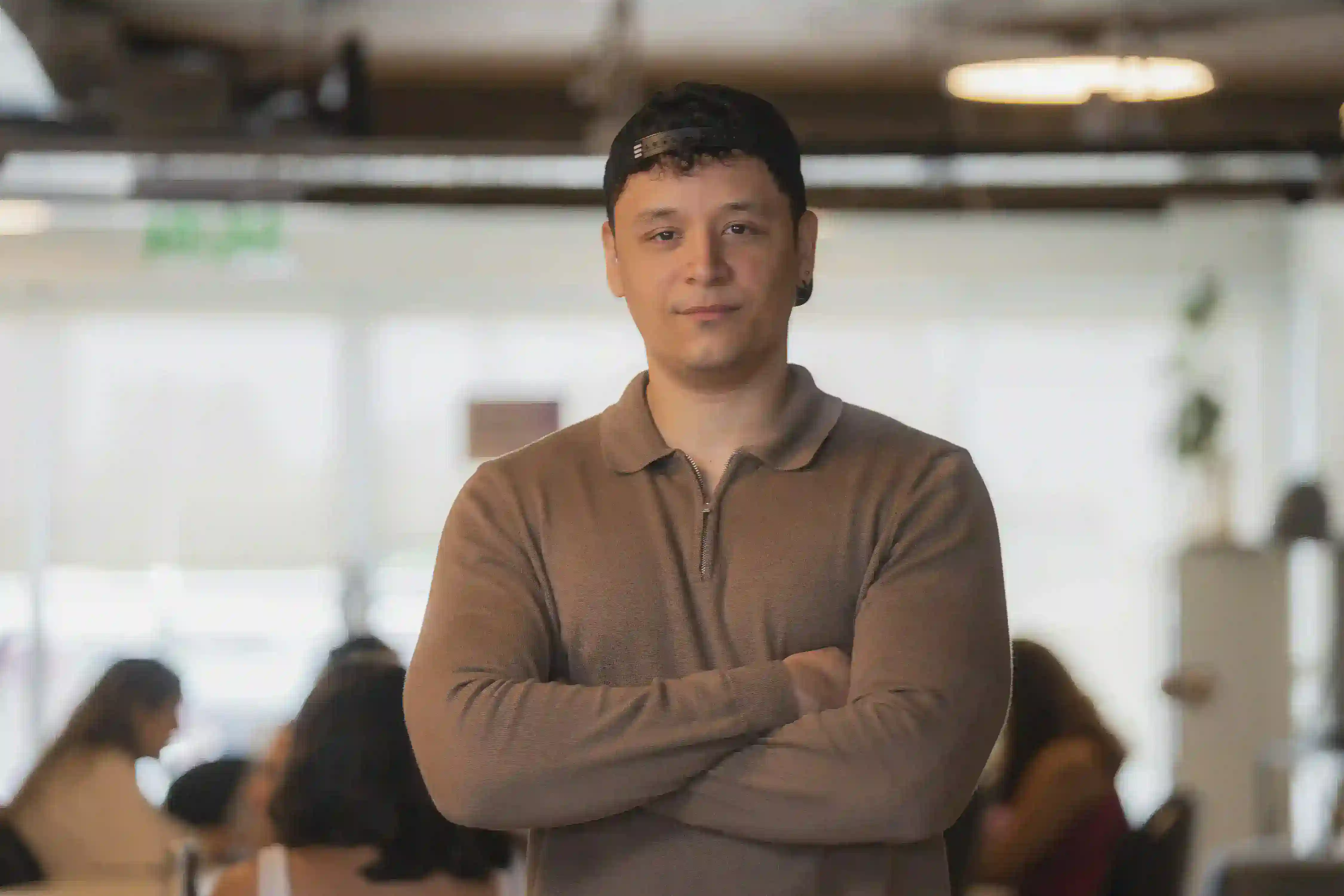 A young man with short, curly hair stands confidently with his arms crossed in a casual environment, wearing a light brown long-sleeve polo shirt and a black cap. The background shows a bright space with blurred figures of people, indicating a social or professional setting.