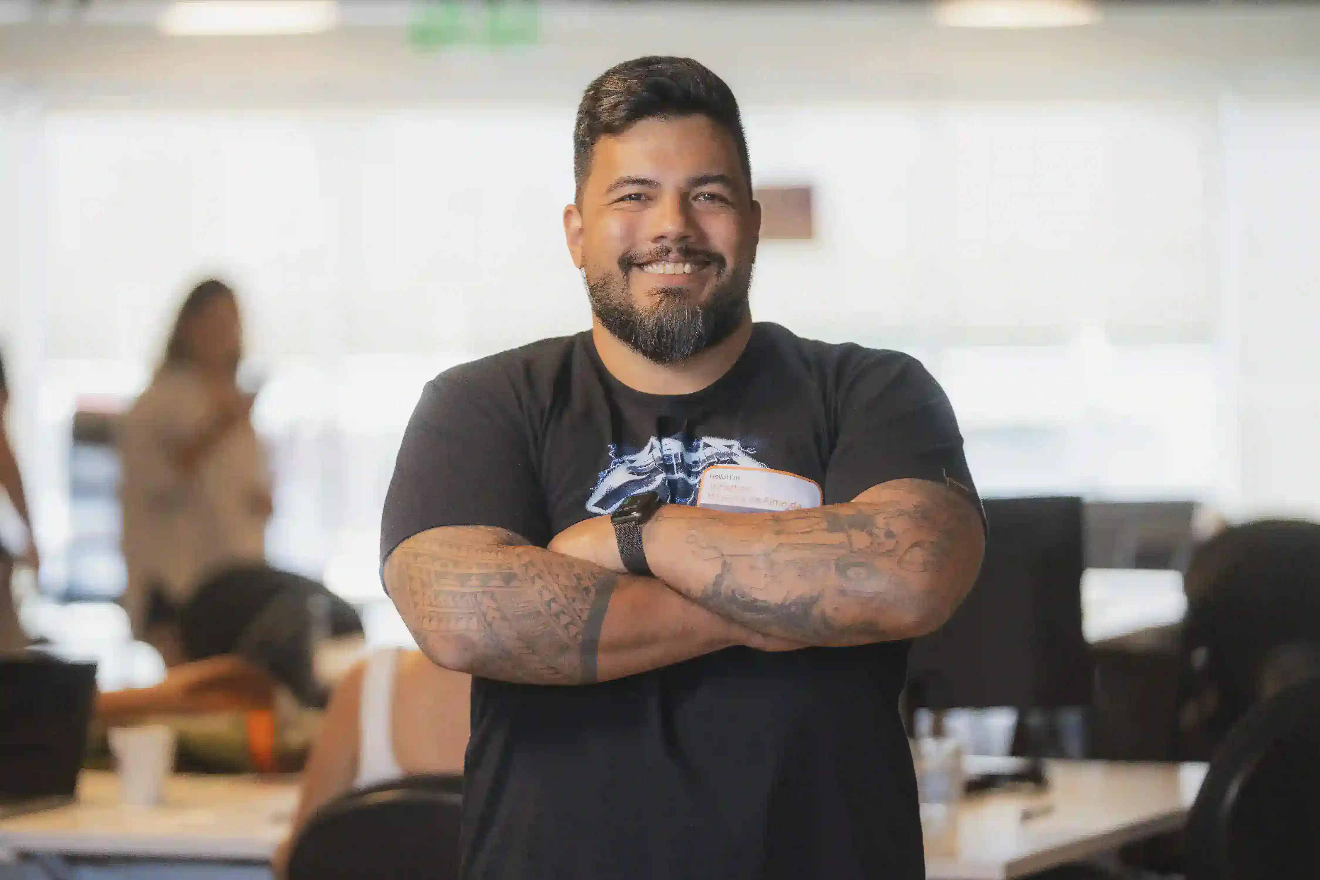 Smiling man with crossed arms and visible tattoos, in an office setting.